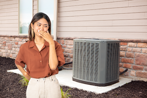 Woman holding her nose while standing next to her outdoor AC unit which smells bad.
