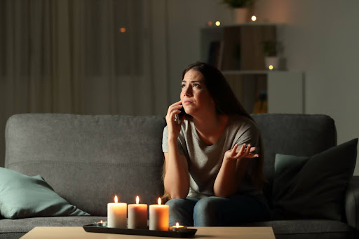 A woman sitting on a couch in a dark room. There are lit candles in front of her on a table as she calls someone on the phone.